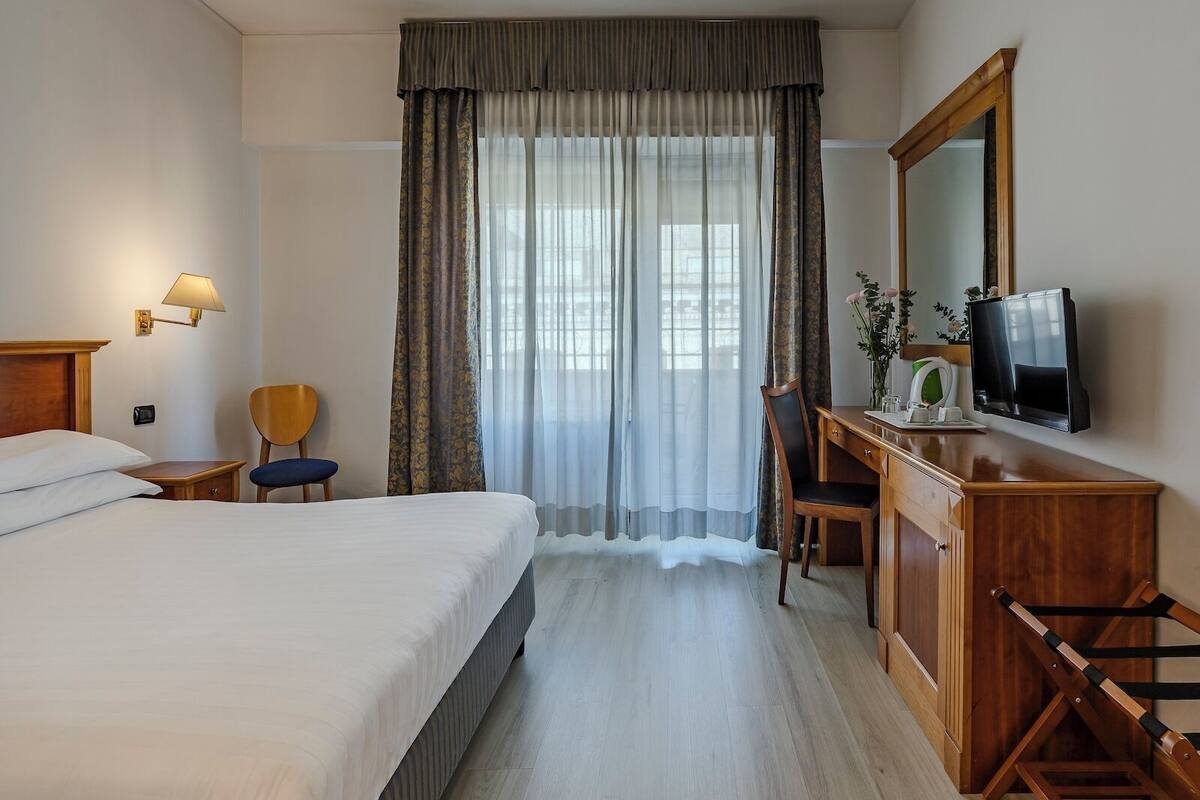 Ein Hotelzimmer mit einem Bett, einem Schreibtisch mit Stuhl, einem Fernseher und einem Fenster mit Vorhängen.