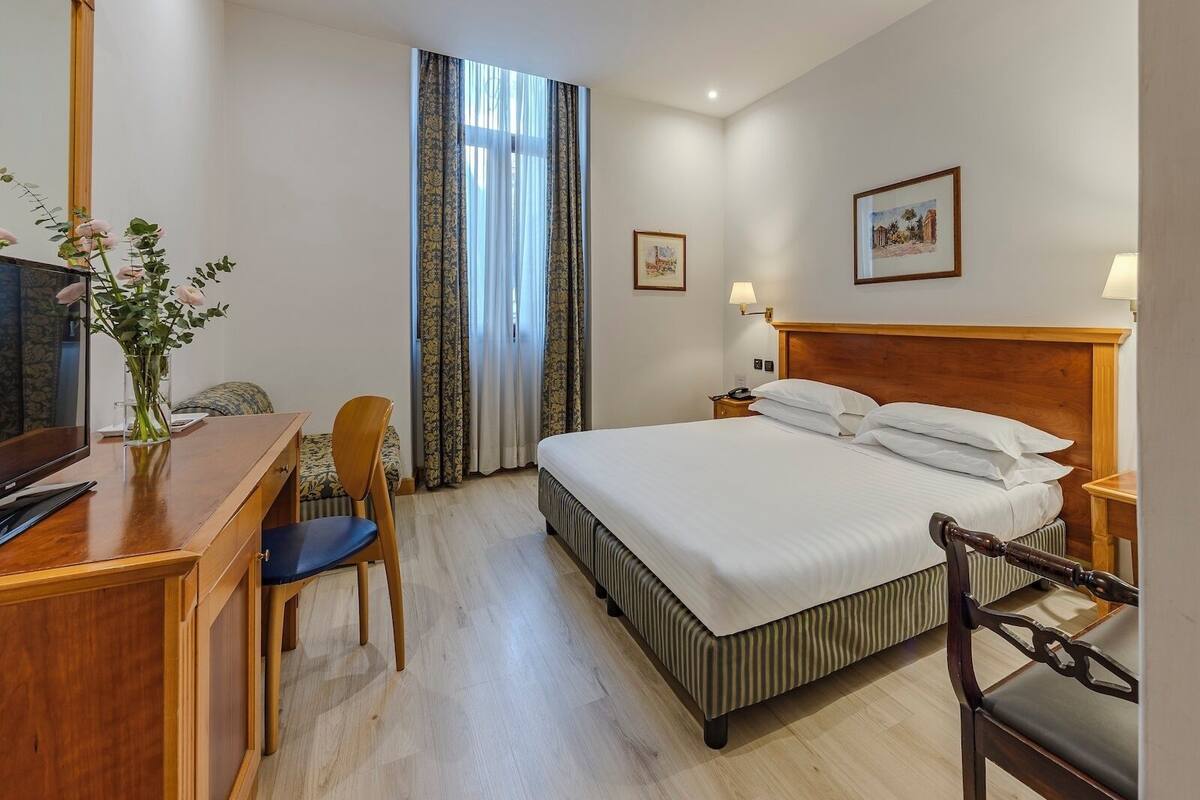 Ein Hotelzimmer mit Bett, Schreibtisch, Stuhl, Fernseher und einem Fenster mit Vorhängen.