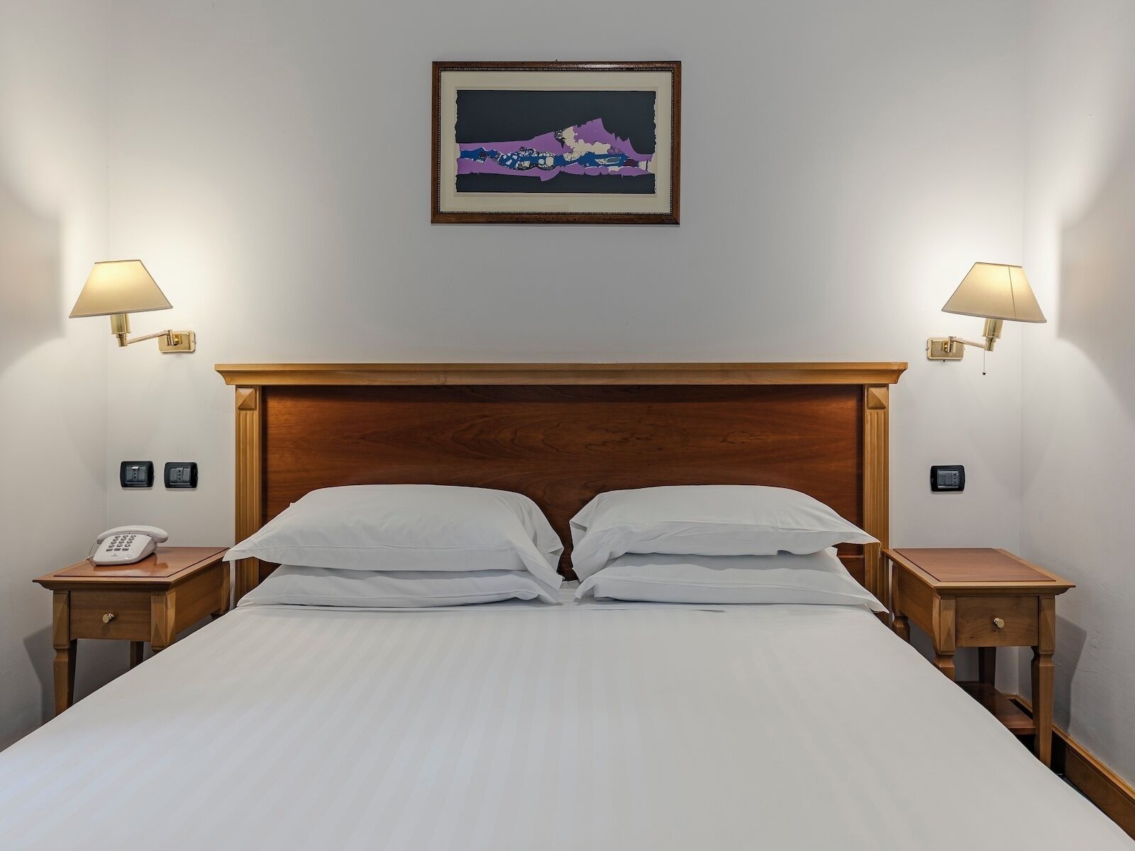 A hotel room with a wooden headboard, a bed with white linens, two bedside tables with lamps, a phone, and a framed picture on the wall.