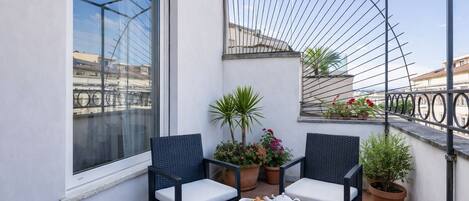 Un balcon avec deux fauteuils en osier, une petite table sur laquelle se trouve un plateau avec de la nourriture et des boissons, des plantes en pot et une vue sur le paysage urbain.