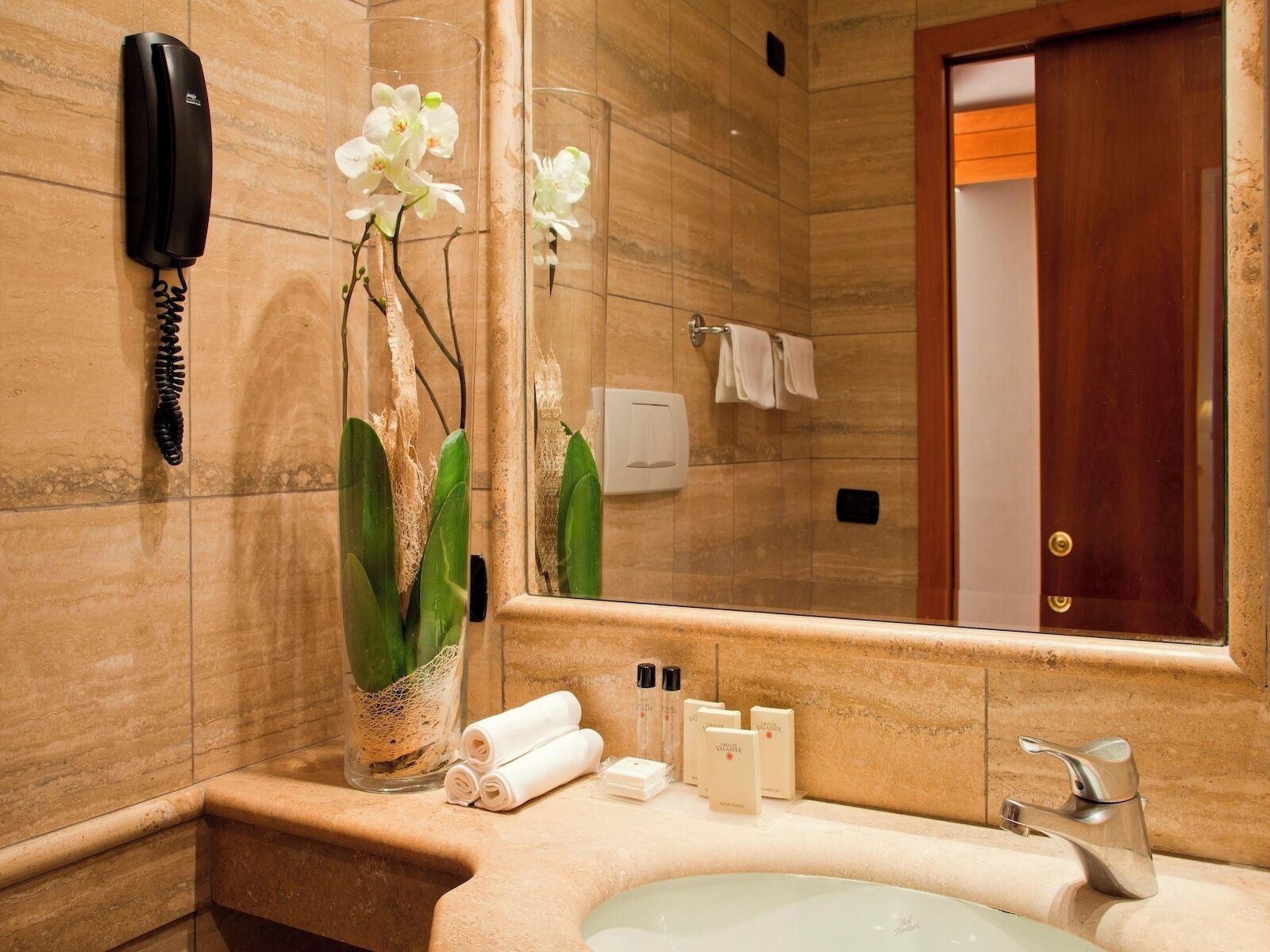 Un baño con pared de mármol, un teléfono, un jarrón con orquídeas, un espejo, un perchero para toallas y artículos de aseo.