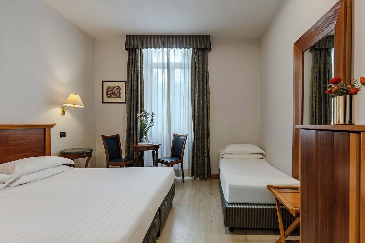 Ein Hotelzimmer mit zwei Betten, einem kleinen Tisch mit Stühlen, einer Lampe, einem Spiegel und einem Fenster mit Vorhängen.