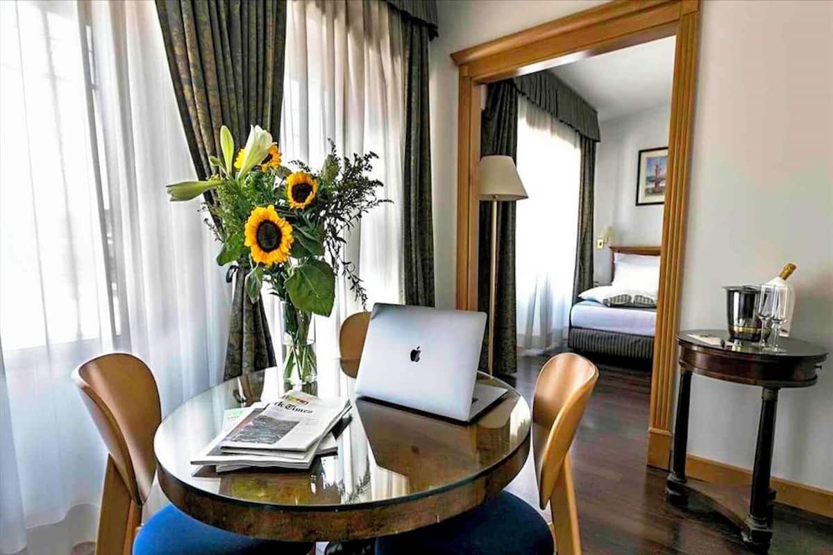 Ein Hotelzimmer mit einem runden Glastisch, einem Laptop, einer Vase mit Sonnenblumen und einem Bett im Hintergrund.