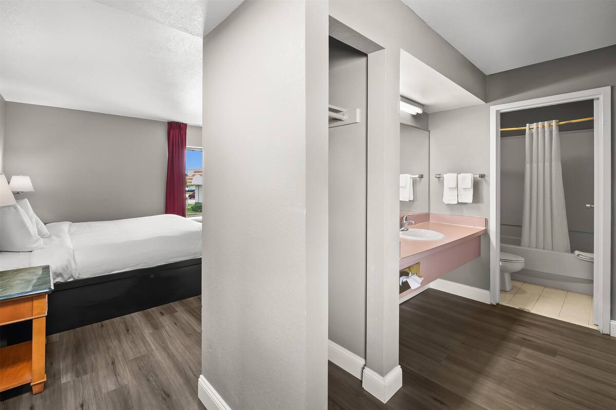 A modern hotel room with a bed, bedside table, and a bathroom with a sink, mirror, and shower.