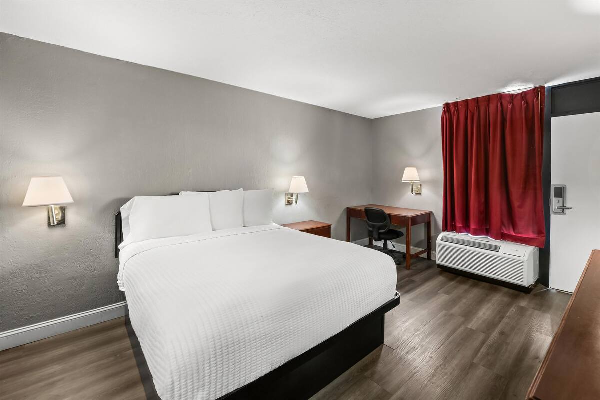 A hotel room with a bed, desk, two wall-mounted lamps, a heater, and a red curtain.
