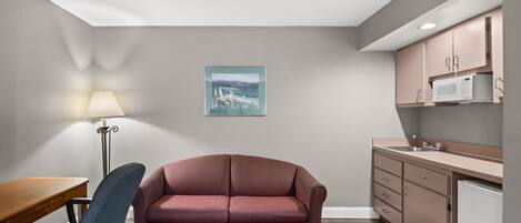 A compact living space with a sofa, a dining table, a lamp, a kitchen area with cabinets and a microwave, and a framed picture on the wall.