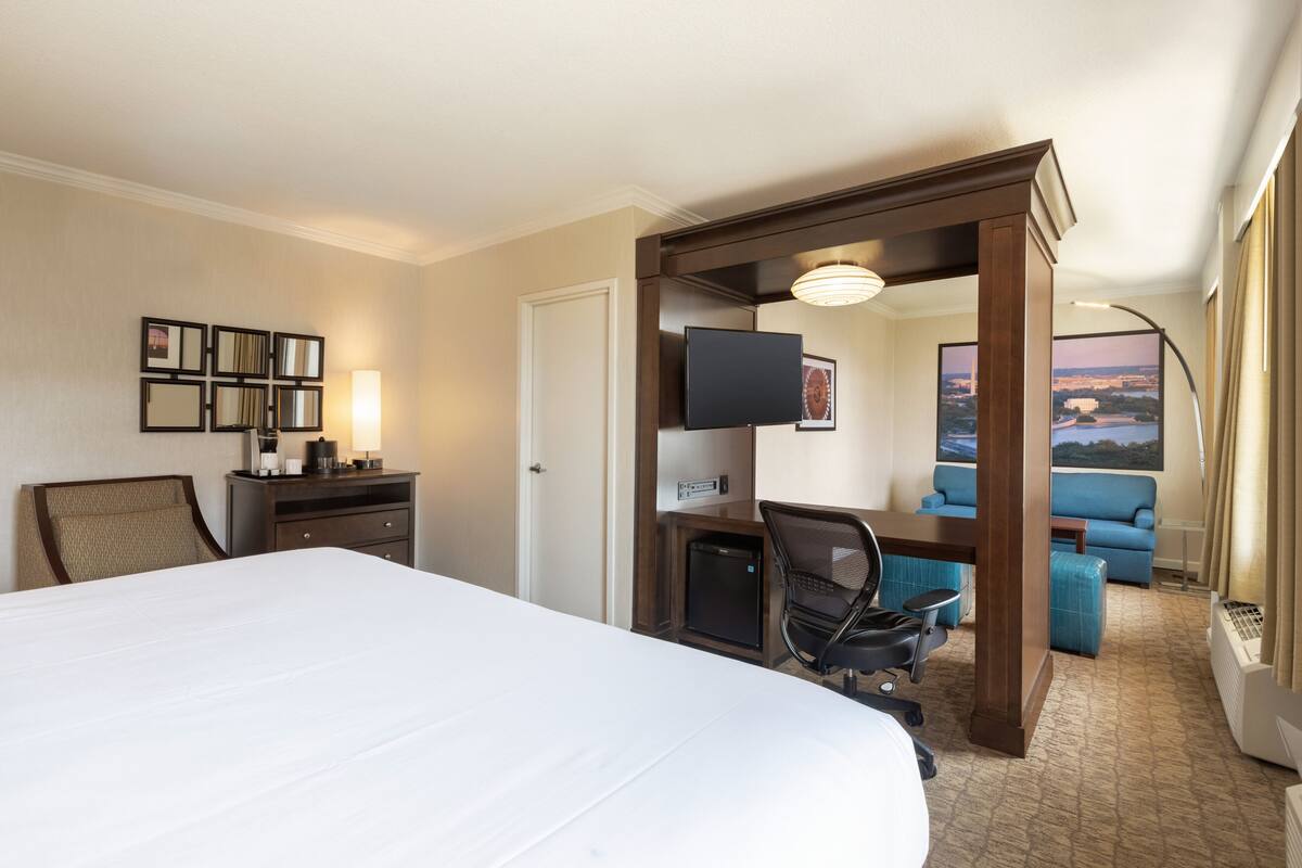 A hotel room with a large bed, a desk with a chair, a television, a small refrigerator, and a view of the city.