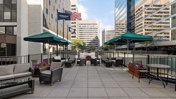 An outdoor seating area with wicker furniture and green umbrellas, surrounded by modern buildings and flags.
