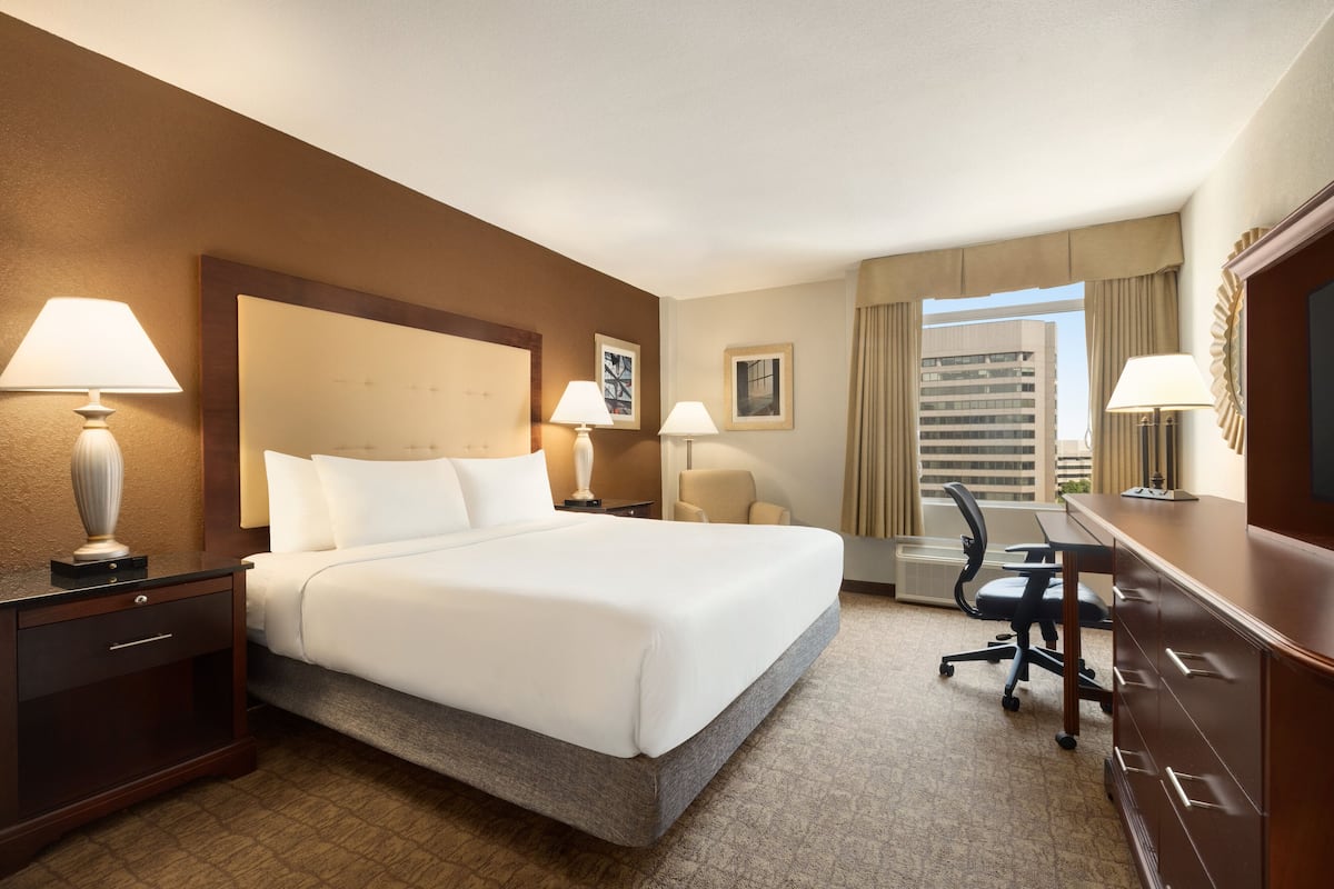 A hotel room with a large bed, two bedside tables with lamps, a desk with a chair, and a view of a building through the window.