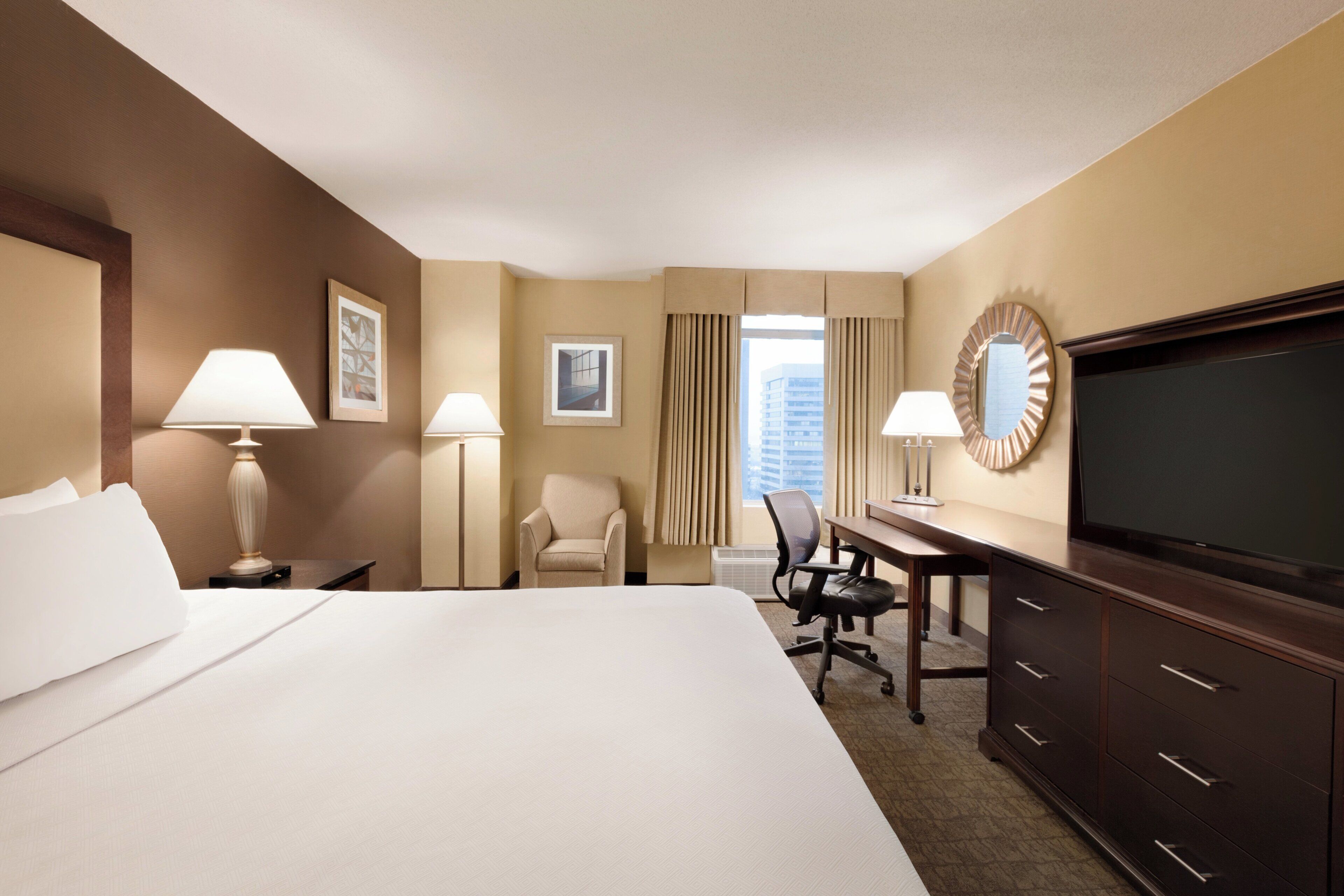 A hotel room with a large bed, a desk, a chair, a television, and two lamps.