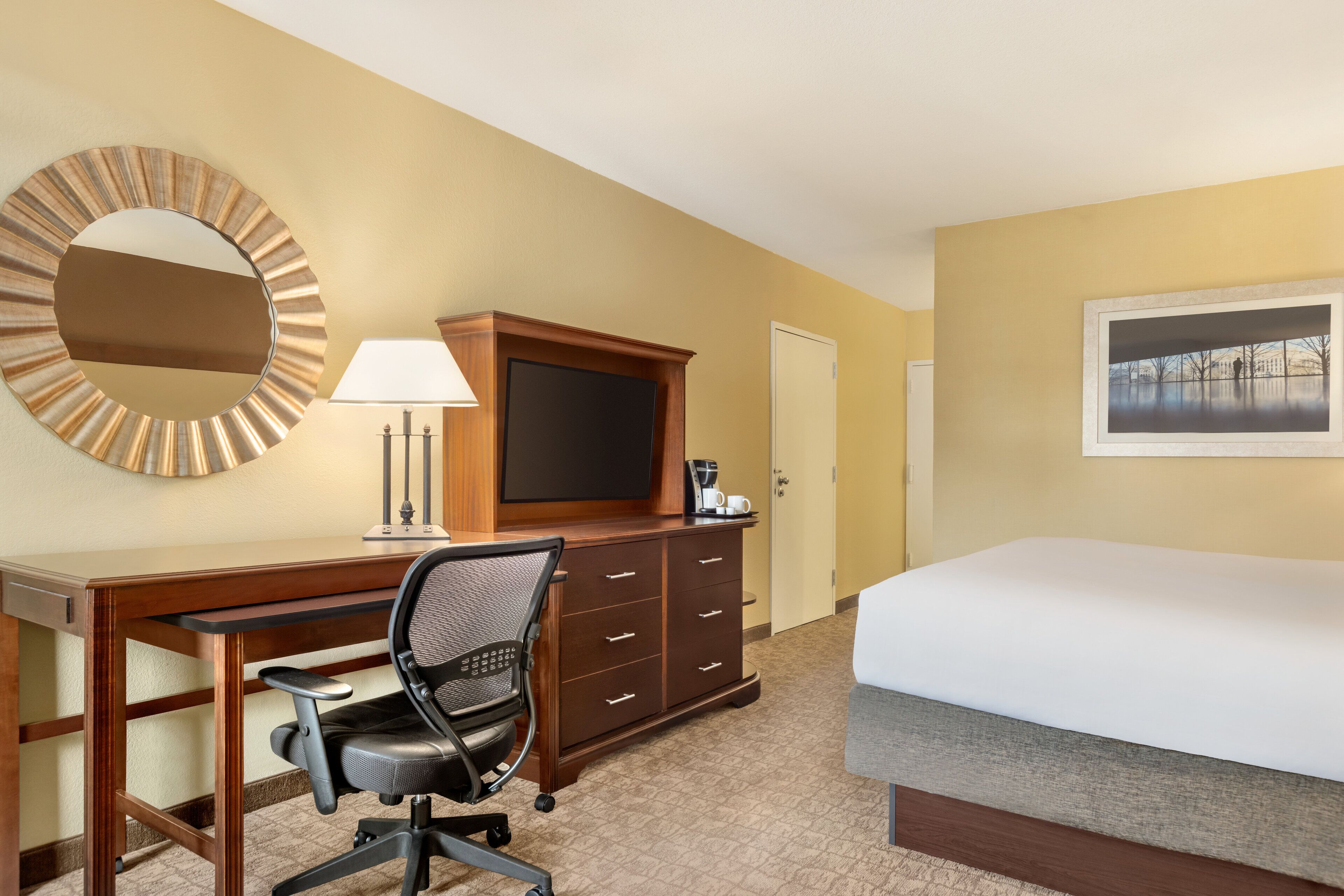 Een hotelkamer met een bed, bureau, stoel, televisie, dressoir, lamp en een schilderij aan de muur.