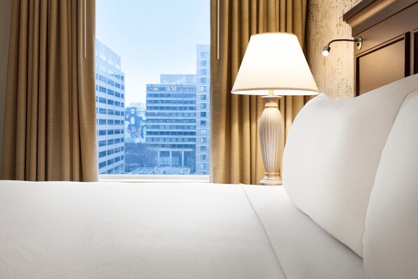 A hotel room with a bed, a lamp, and a view of city buildings through the window. - Crowne Plaza Crystal City-Washington, D.C. by IHG (Arlington)
