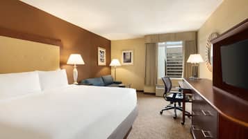 A hotel room with a large bed, a desk, two chairs, a TV, and a window with curtains.