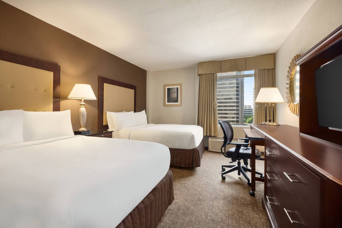A hotel room with two beds, a desk, a chair, a TV, and a window with curtains.