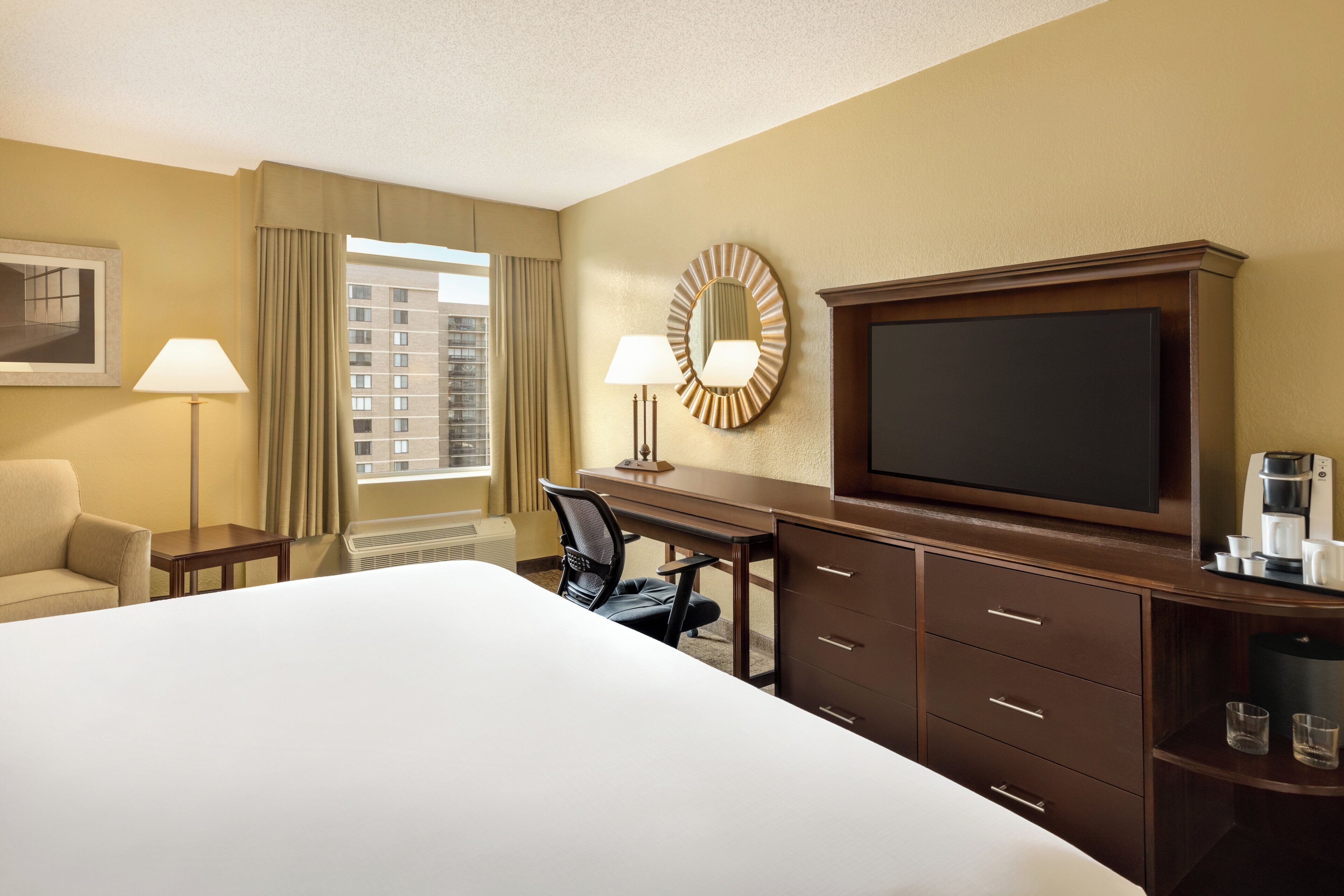 A hotel room with a large bed, a desk with a chair, a television, a coffee maker, and a window with curtains.