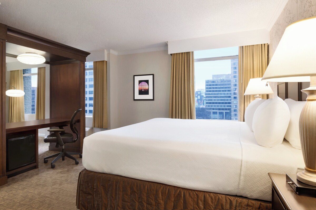 A hotel room with a large bed, a desk, a chair, a lamp, and a framed picture on the wall.