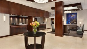 Eine Hotelhalle mit einer Rezeption, einem runden Tisch mit Blumenvase, einer Sitzecke mit Sofa und Sesseln sowie einem großen Wandbild.