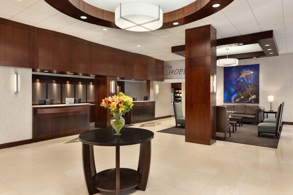 A hotel lobby with a reception desk, a round table with a vase of flowers, a seating area with a sofa and chairs, and a large wall mural. - Crowne Plaza Crystal City-Washington, D.C. by IHG (Arlington)