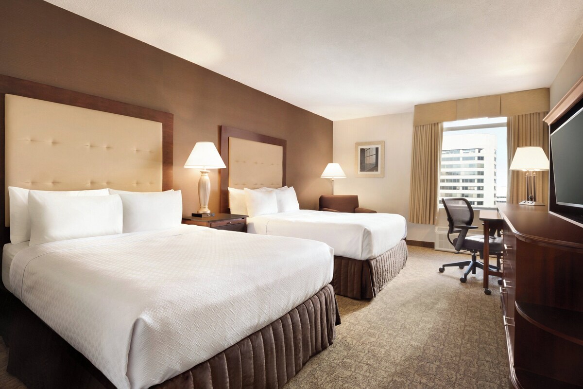A hotel room with two beds, a desk, a chair, lamps, a television, and a window with curtains.