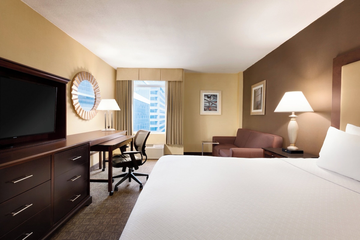 Quarto de hotel com cama, escrivaninha com cadeira, televisão, abajur e janela com cortinas.