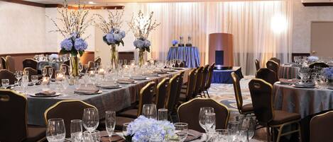 Een feestzaal met ronde tafels gedekt voor een diner, met transparant glaswerk en bloemstukken als decoratie.