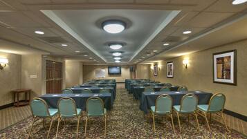 Un salón de conferencias con filas de mesas y sillas, una alfombra estampada y una pantalla de televisión al frente.