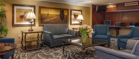 Un vestíbulo de hotel con un sofá gris, sillones azules, una mesa de centro con un jarrón de flores rosadas, obras de arte enmarcadas en las paredes y un televisor sobre un mueble.
