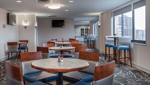 A hotel dining area with round tables and cushioned chairs, a flat-screen TV mounted on the wall, and large windows offering a view of the cityscape. - Arlington Court Suites, a Clarion Collection Hotel (Arlington)