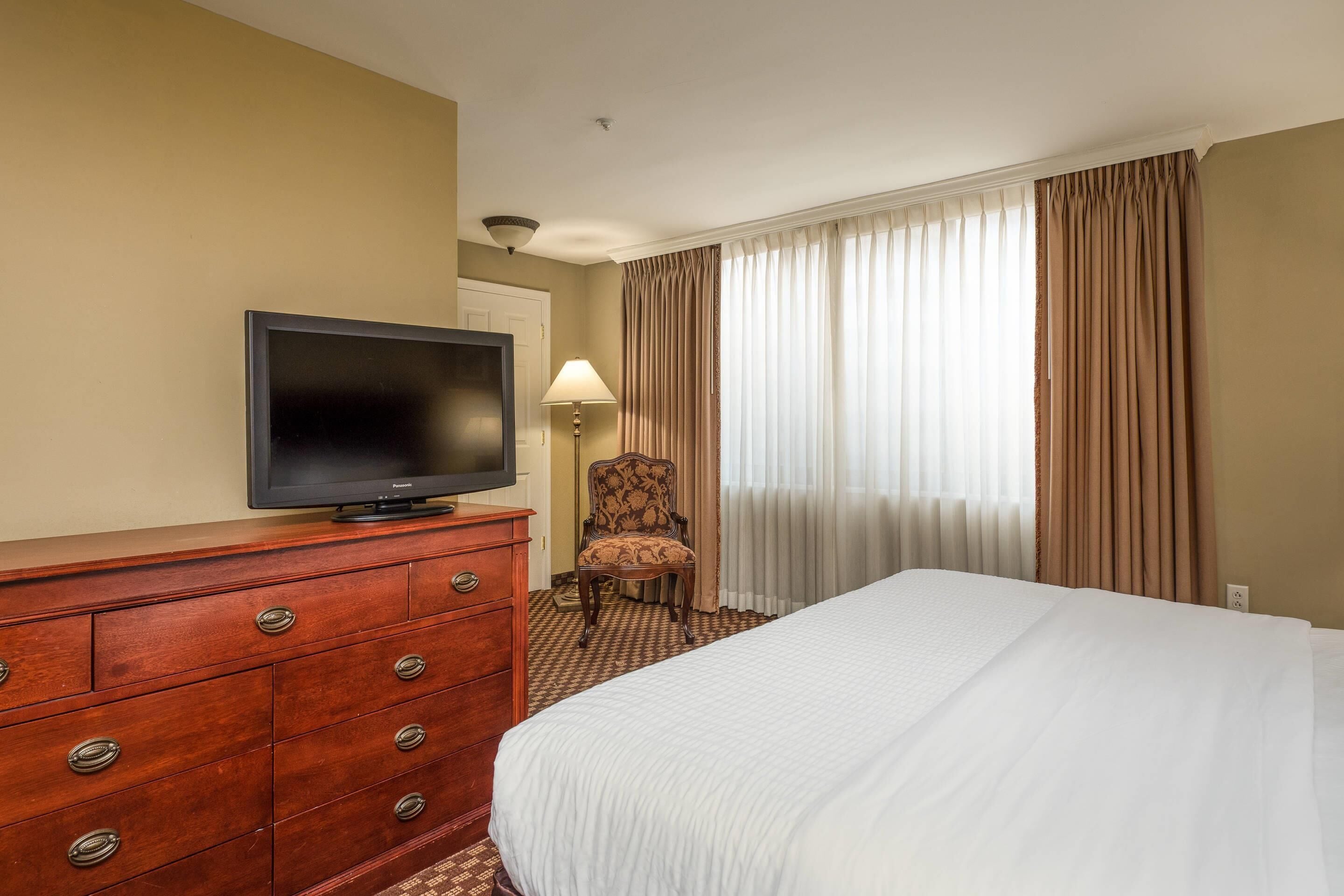 Une chambre d’hôtel avec un lit, un téléviseur posé sur une commode en bois, une chaise et une fenêtre avec des rideaux.