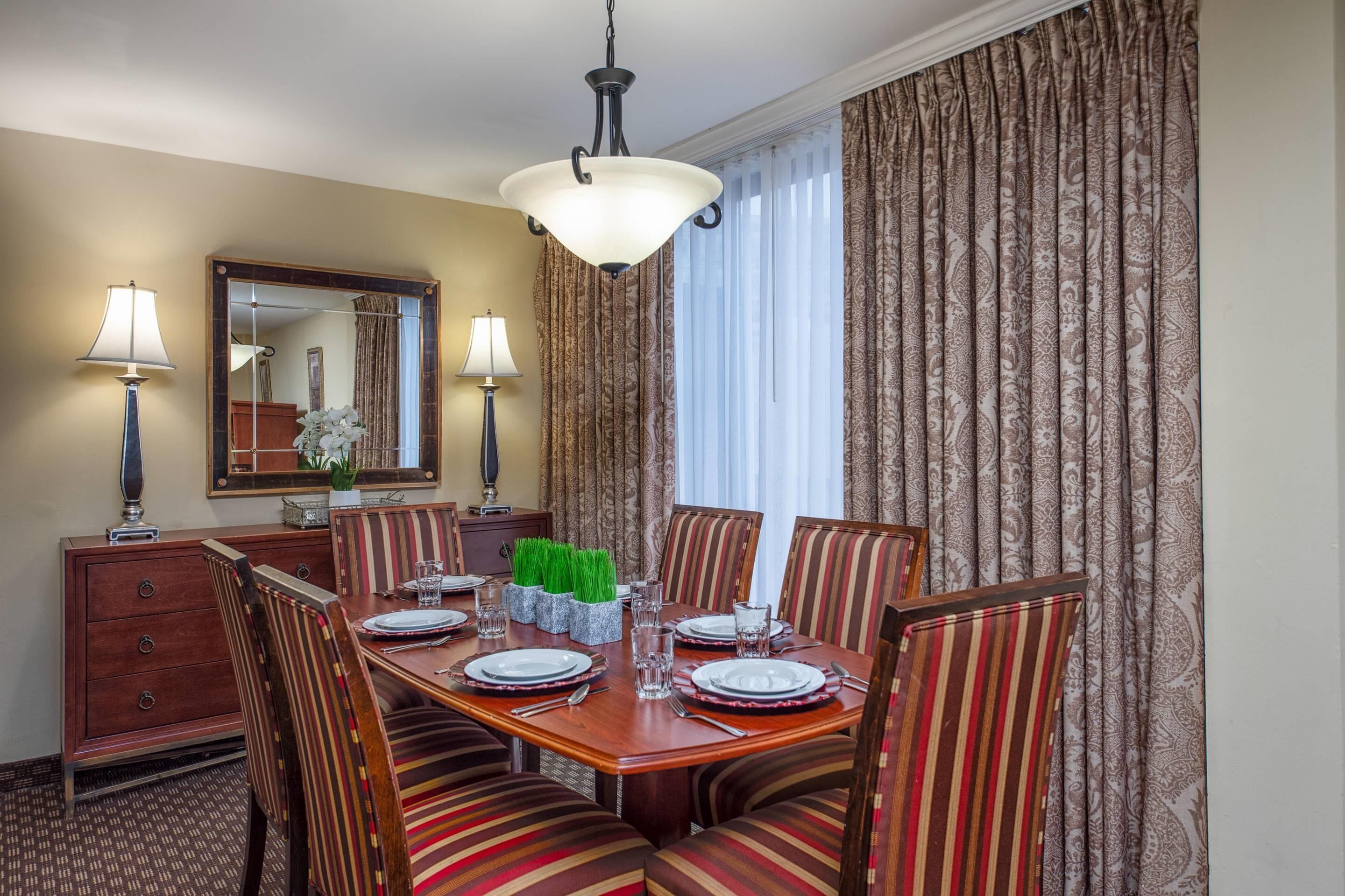 Un comedor con una mesa de madera dispuesta para cuatro personas, sillas a rayas, dos lámparas, un espejo y una ventana con cortinas estampadas.