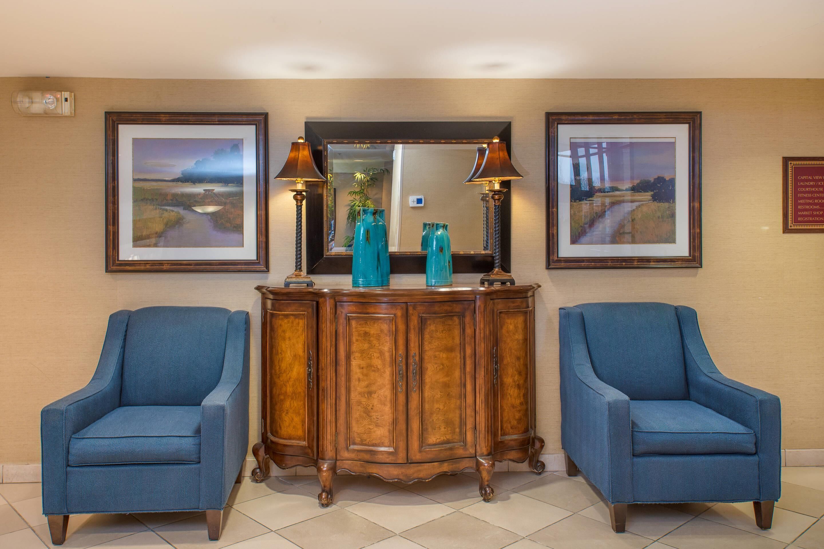 A lobby with two blue armchairs, a wooden console table with decorative items, framed landscape paintings, and a mirror above the console.