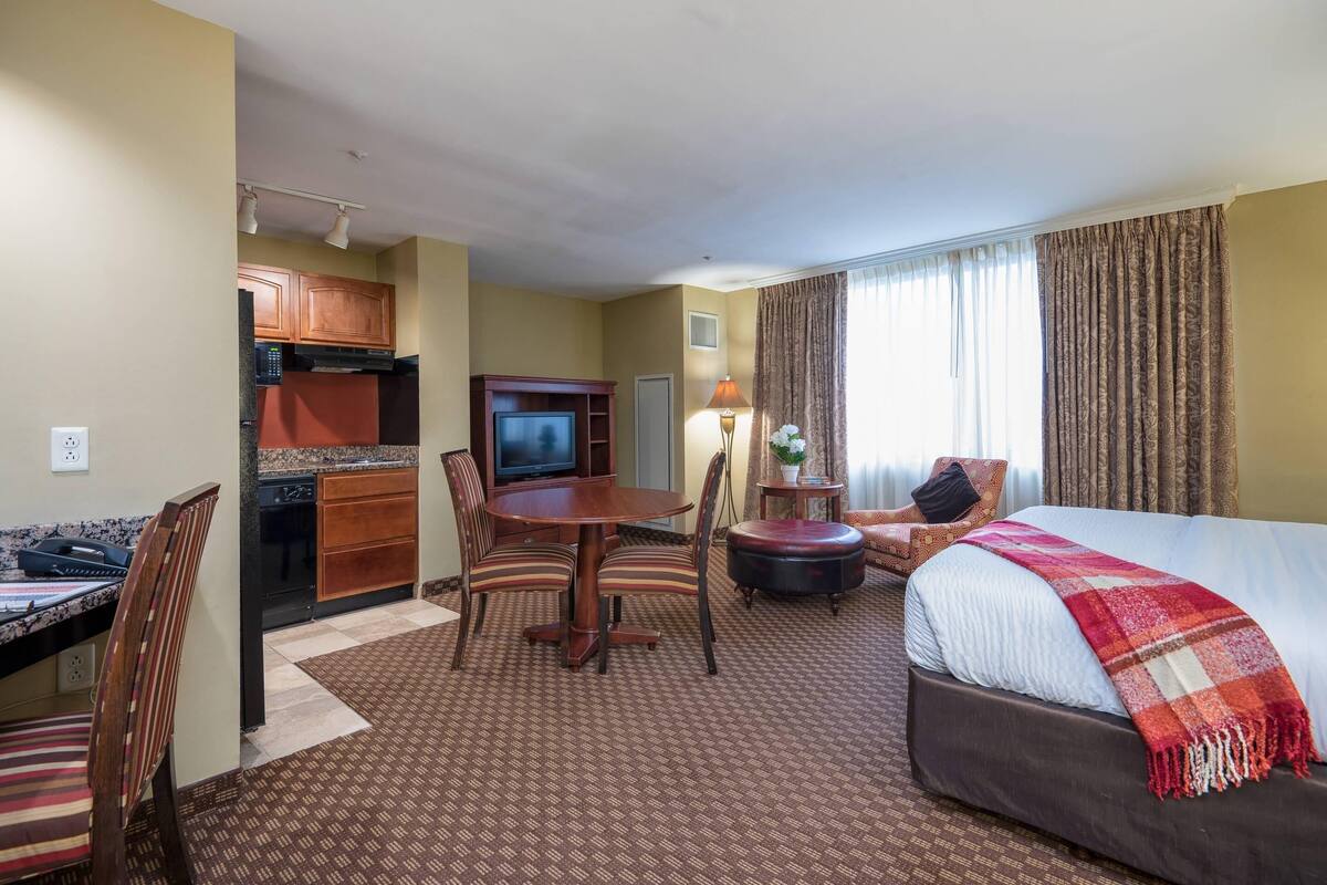 A hotel room with a bed, a desk with a chair, a dining table with chairs, a television, and a kitchen area.