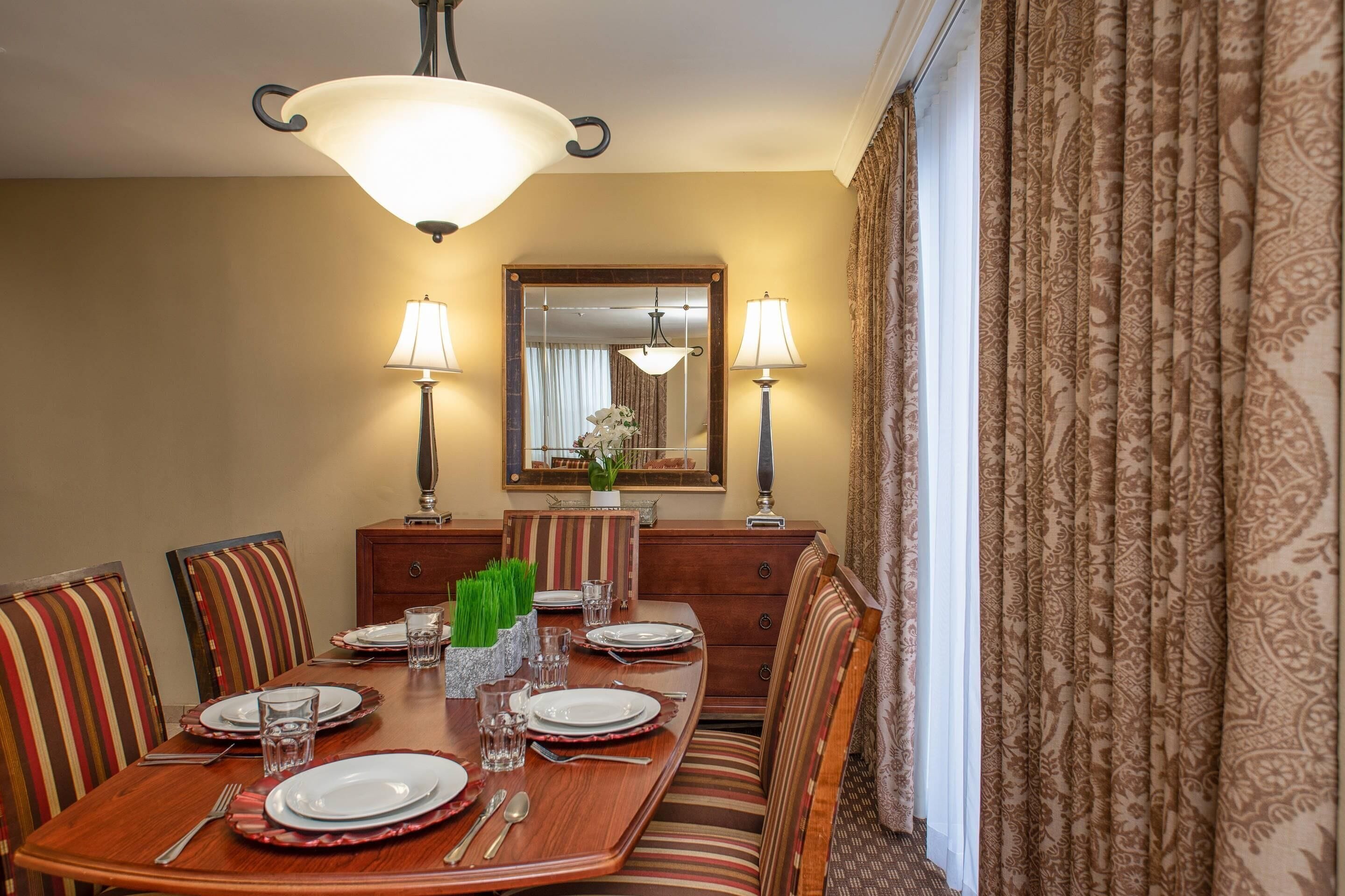 Un comedor con una mesa de madera dispuesta para cuatro personas, flanqueada por sillas a rayas, un espejo sobre la mesa y cortinas estampadas en la ventana.