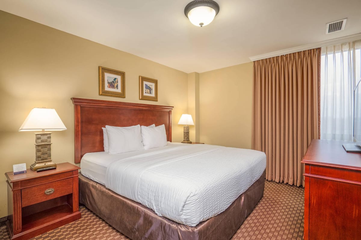 A hotel room with a large bed, two bedside tables with lamps, a wooden headboard, and a view of the outside through curtains.