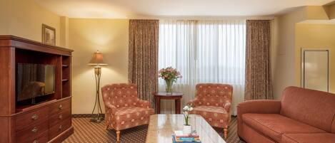 A hotel room with a wooden cabinet, two patterned armchairs, a sofa, a coffee table with books, a vase with flowers, and a large window with curtains.