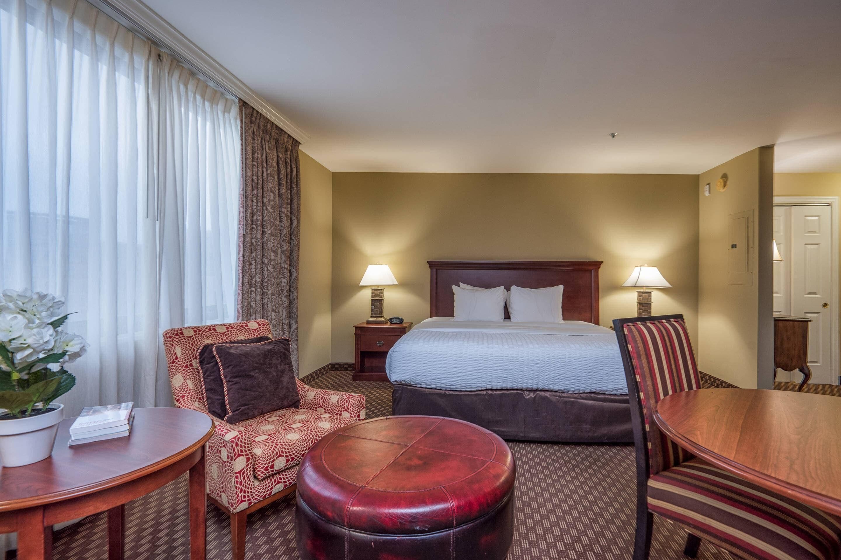 Habitación de hotel con una cama, dos lámparas de noche, una mesa de madera, una silla tapizada en rojo, un sillón estampado y un otoman redondito.