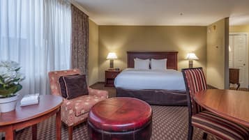 Habitación de hotel con una cama, dos lámparas de noche, una mesa de madera, una silla tapizada en rojo, un sillón estampado y un otoman redondito.
