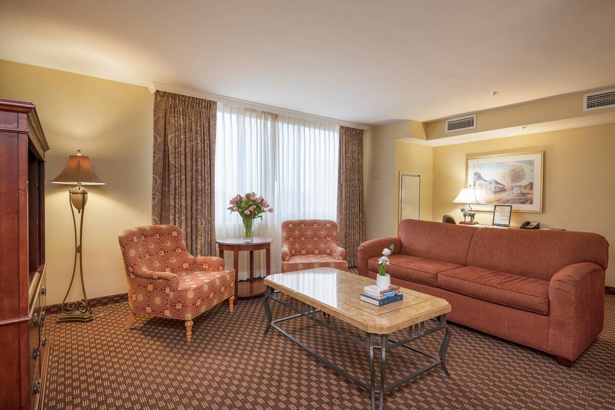 A hotel room with a patterned carpet, two patterned armchairs, a sofa, a small table with books, a vase with flowers, and a painting on the wall.