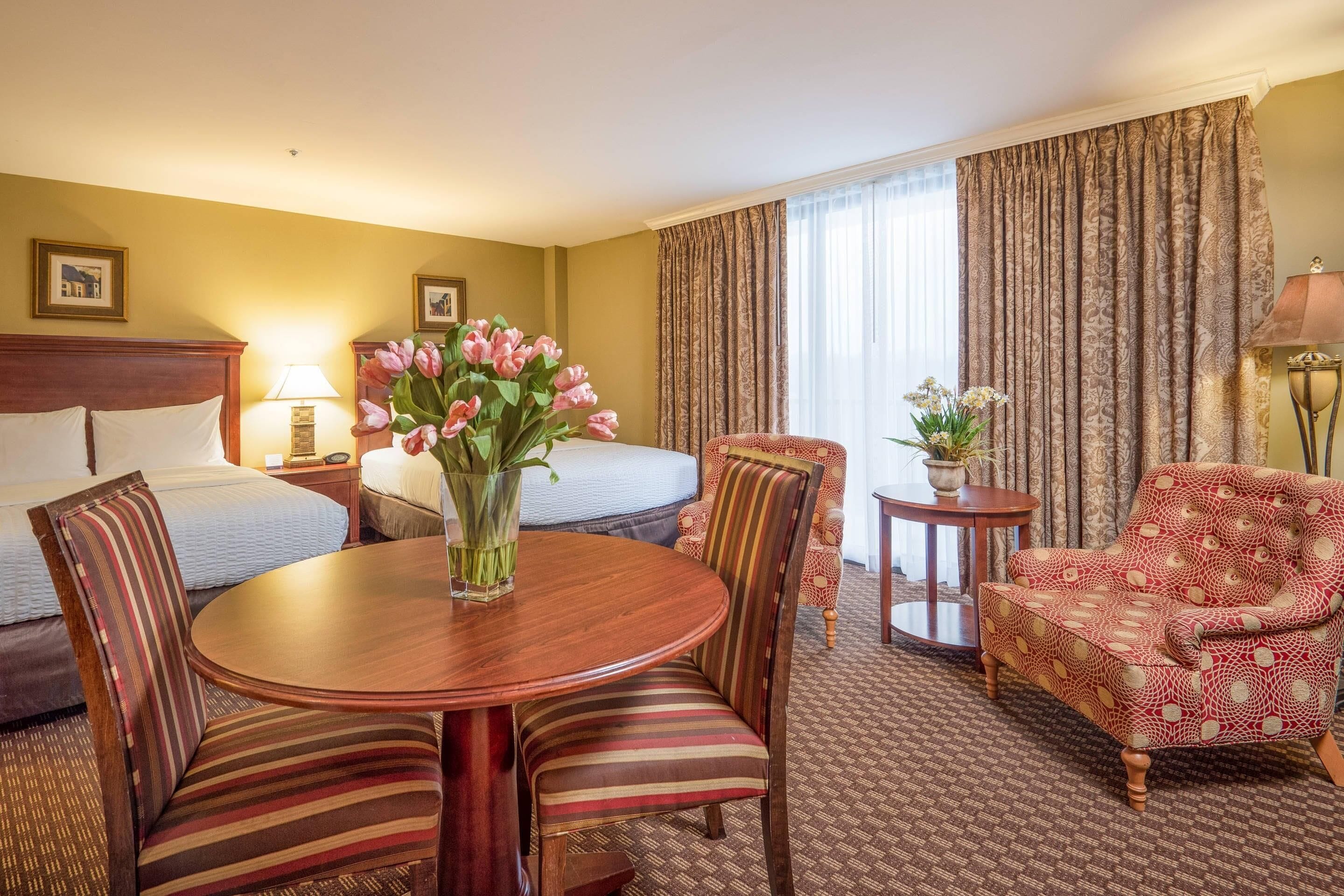A hotel room with a bed, a dining table with chairs, a small round table with a lamp, a patterned armchair, and a vase with flowers.