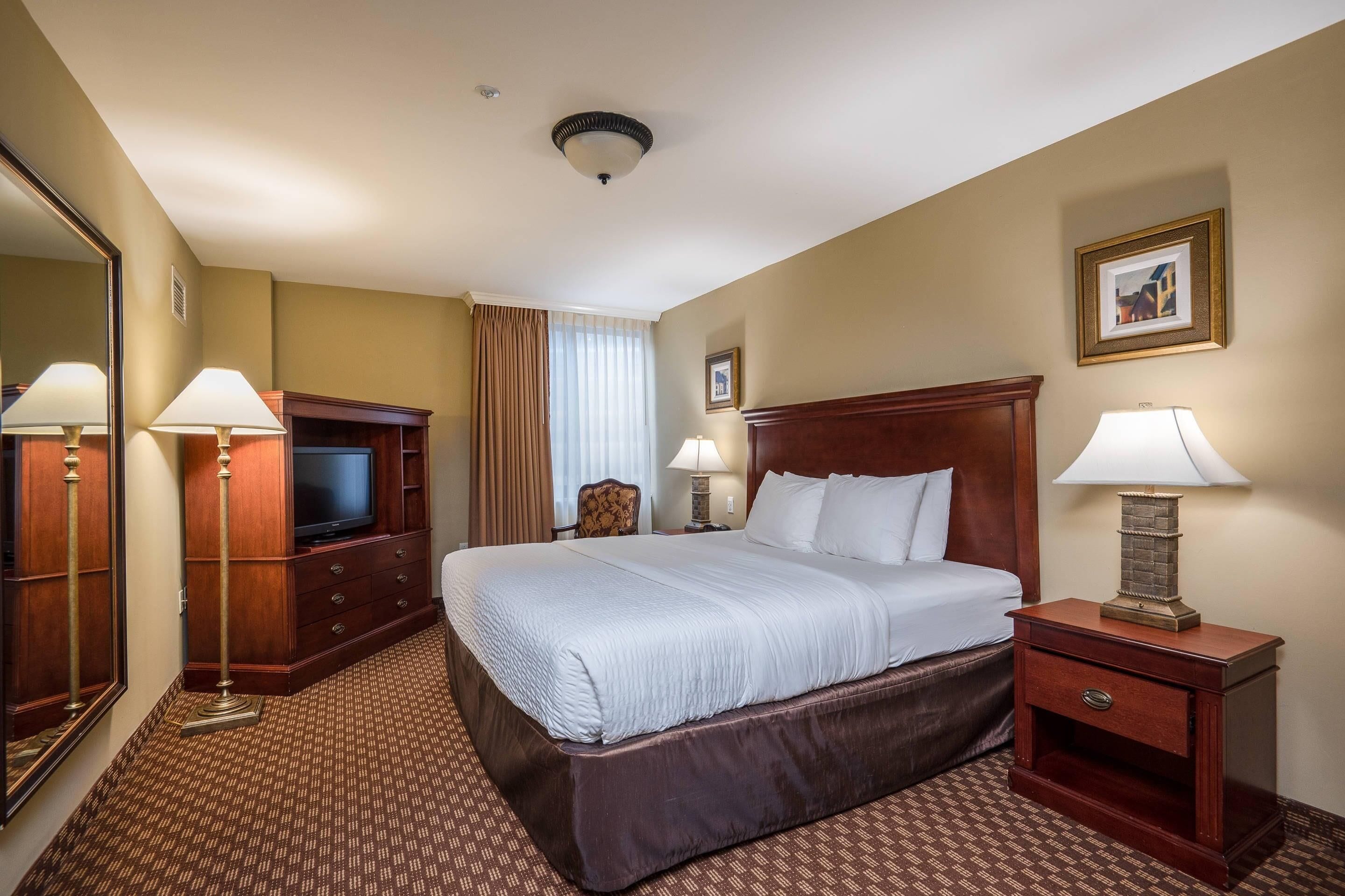 Une chambre d’hôtel avec un grand lit, deux tables de chevet avec des lampes, une télévision, un miroir et un tableau encadré au mur.