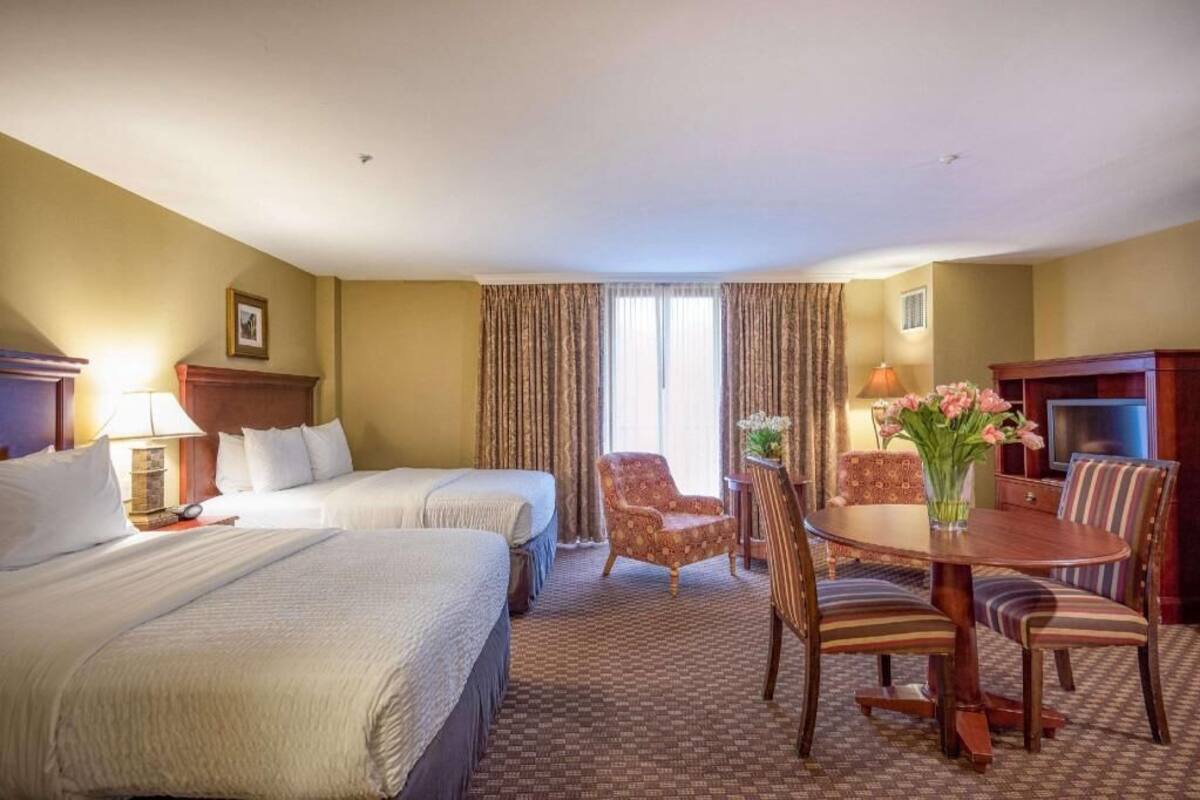 A hotel room with two beds, a dining table with chairs, a TV, and a window with curtains.
