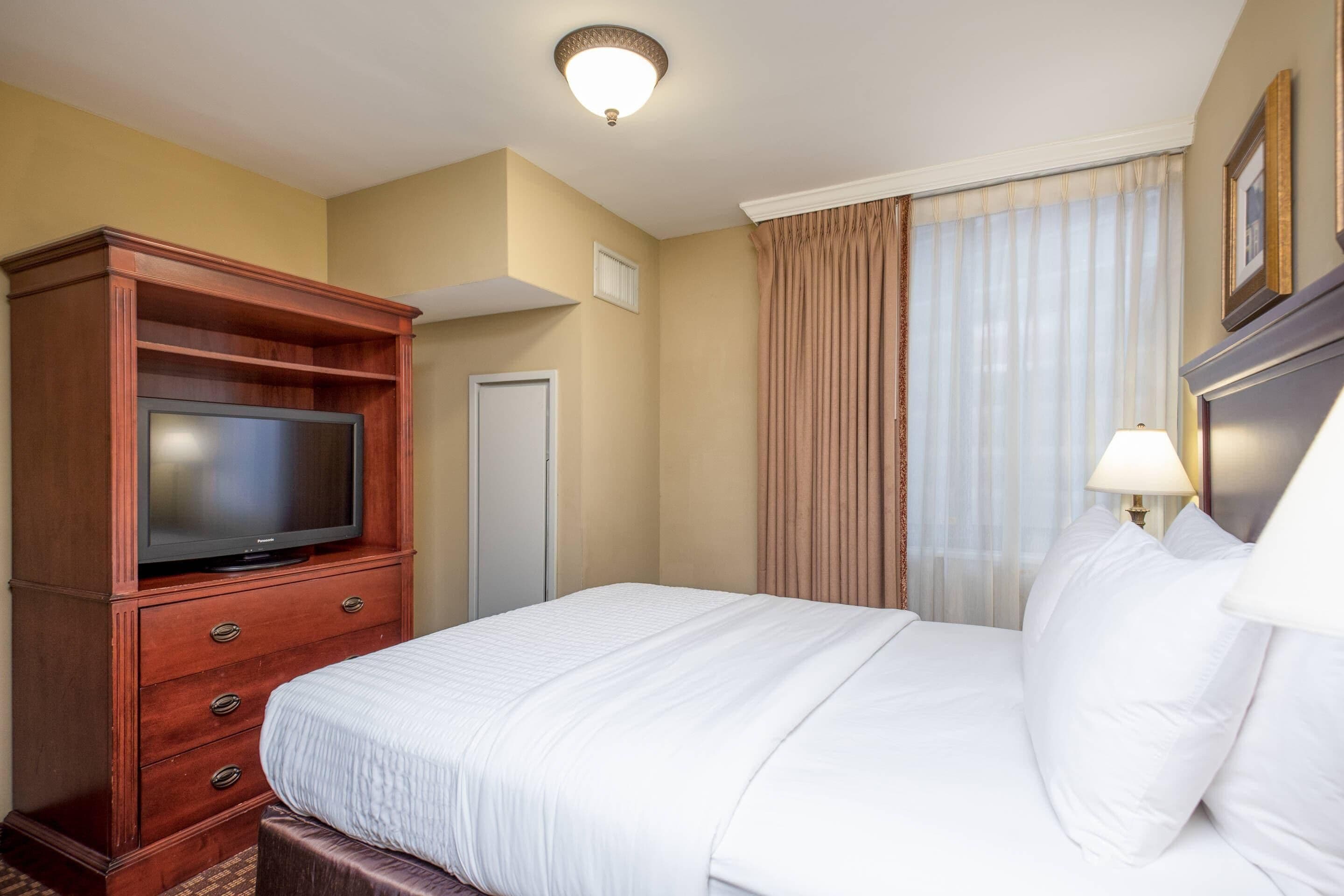 A hotel room with a large bed, a wooden dresser with a flat-screen TV, a door, and a window with curtains.