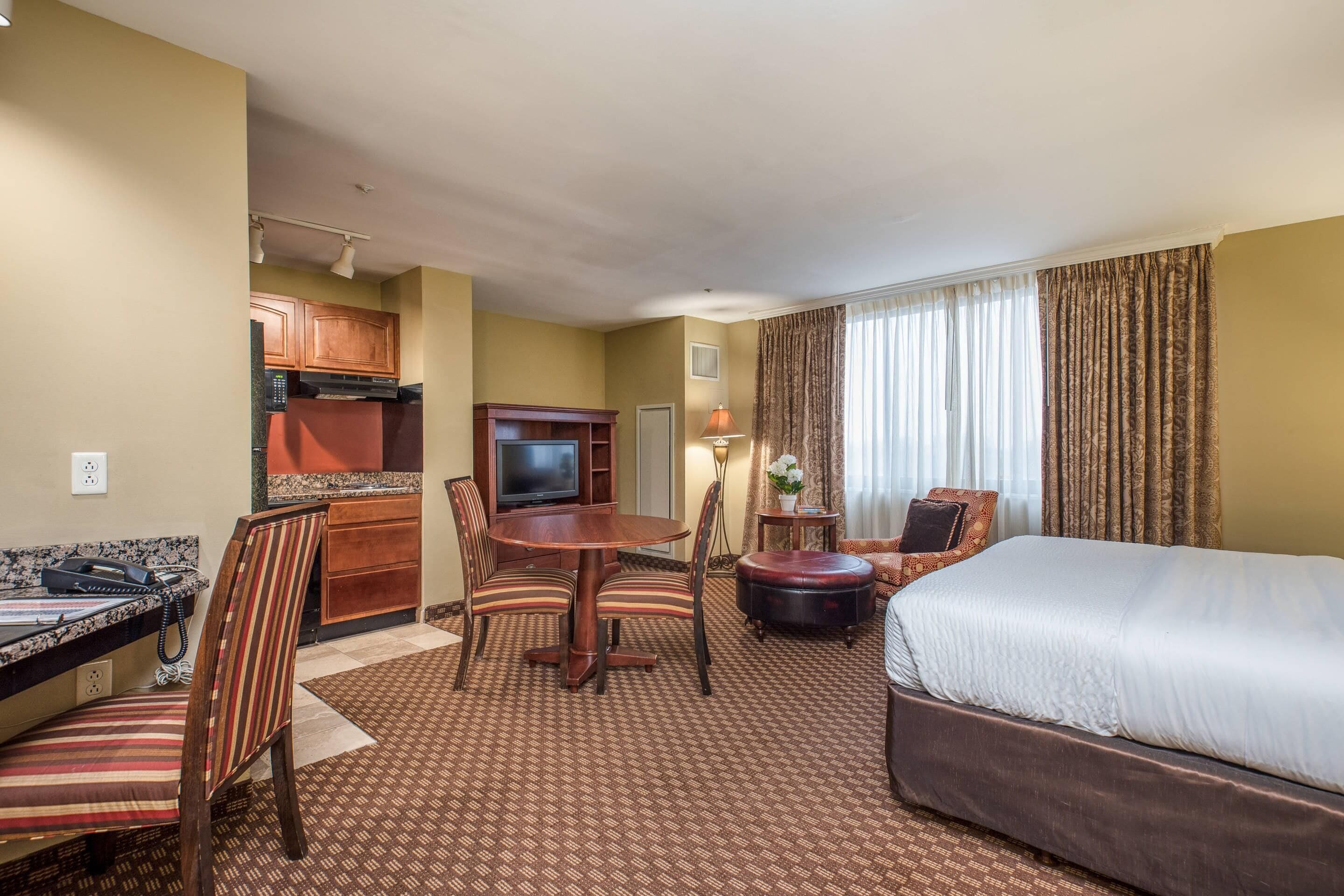 Una habitación de hotel con cama, una mesa de comedor con sillas, televisión, una mesita con lámpara y una zona de cocina.