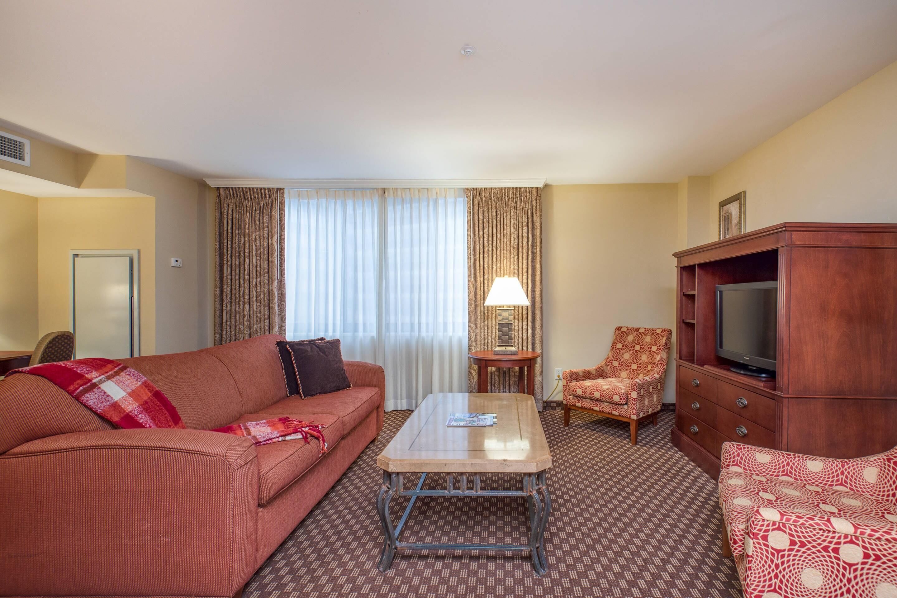 Habitación de hotel con un sofá rojo, una mesa de centro de madera, un mueble para televisor y una alfombra estampada.
