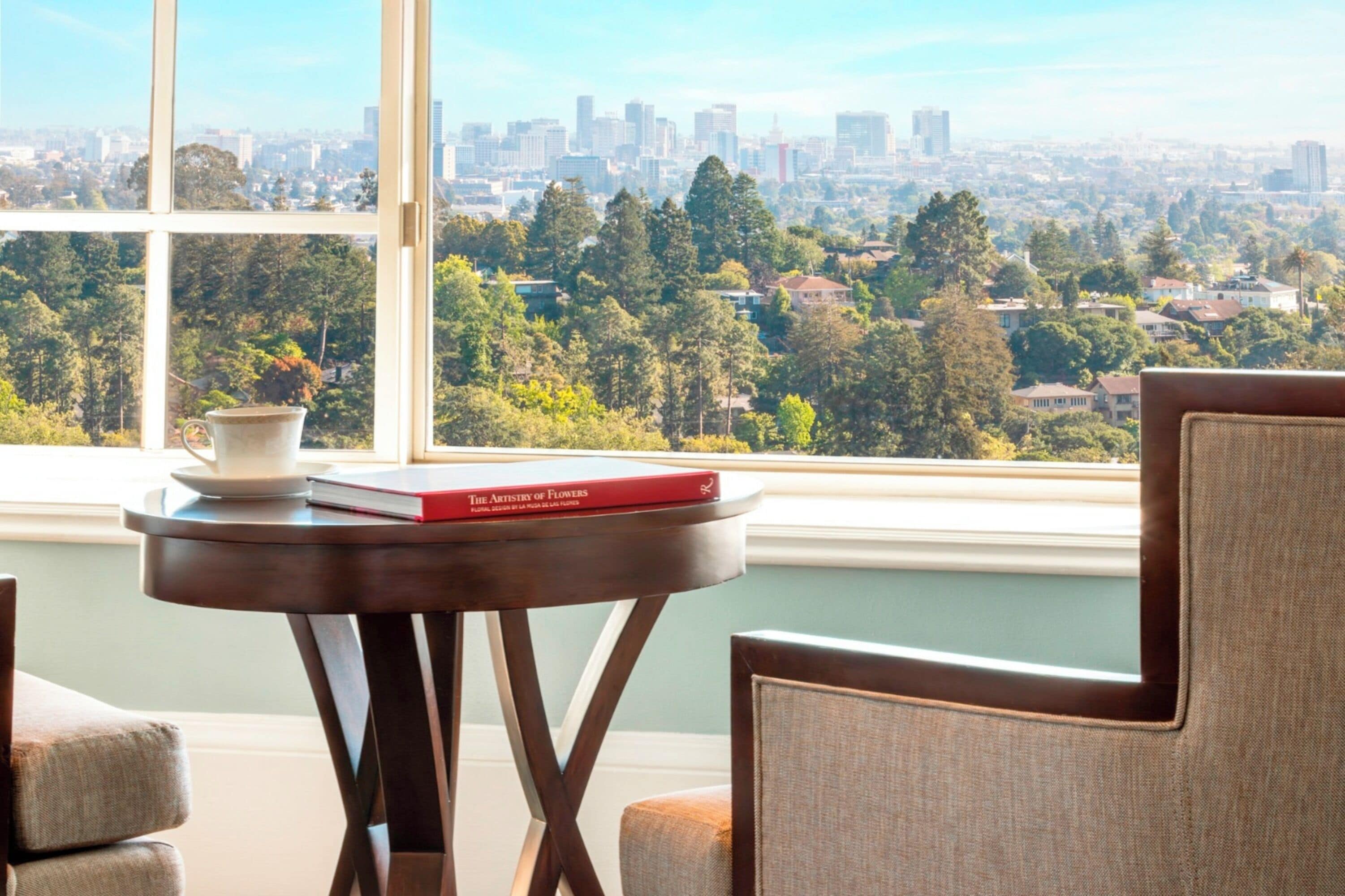 Una habitación con un ventanal grande que ofrece vistas de la ciudad, una mesita redonda con un libro y una taza, y una silla.