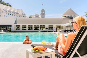 Un área de comedor junto a la piscina, con una mesa preparada para dos, una piscina y un edificio al fondo con una torre y cúpula distintivas.