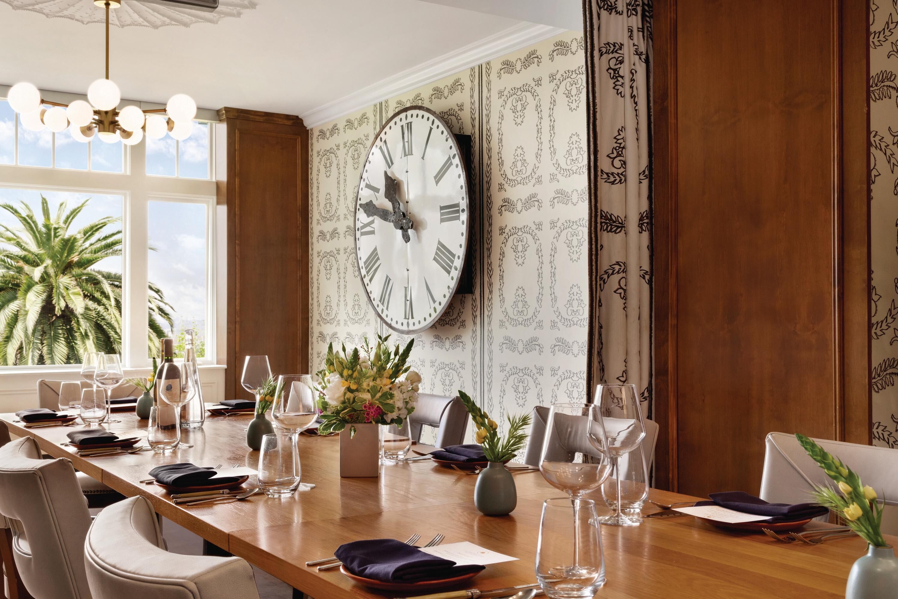 Un comedor con un reloj de pared grande, una mesa larga dispuesta para una comida, y una ventana que ofrece vistas a palmeras.
