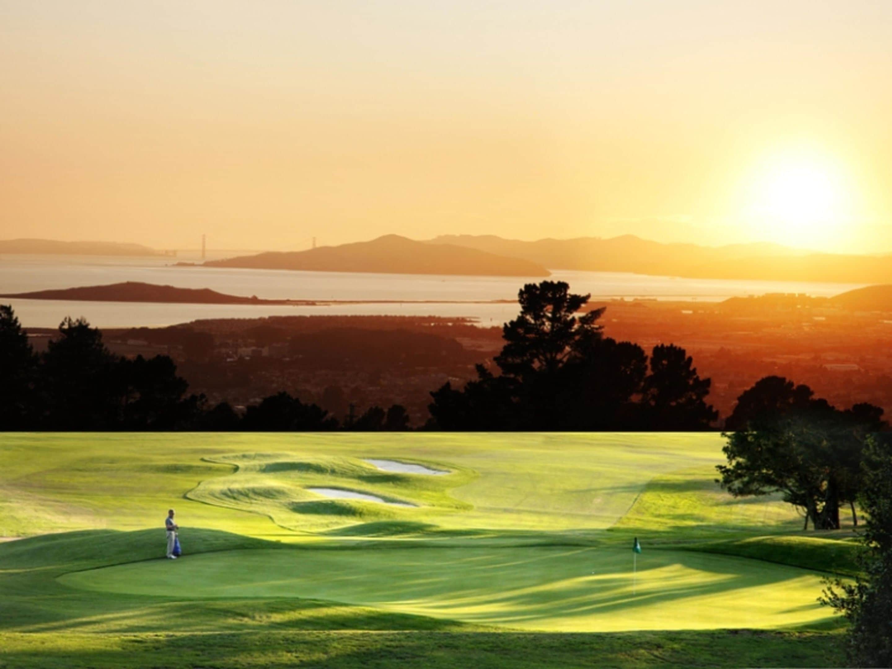A golf course with a person standing near the hole, a flag, and a sunset in the background.