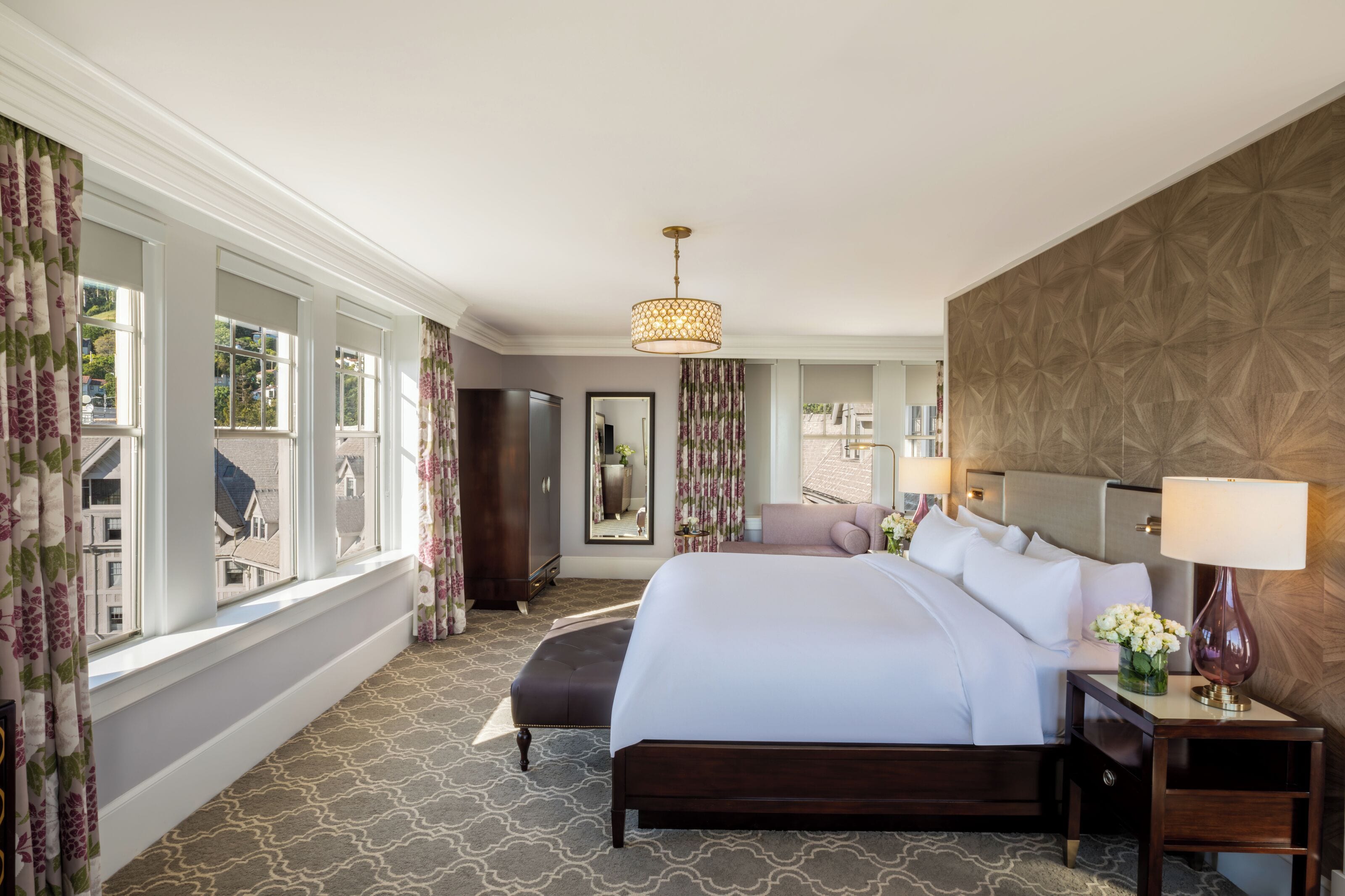 A hotel room with a large bed, a nightstand, a lamp, a mirror, and a view of the outside.