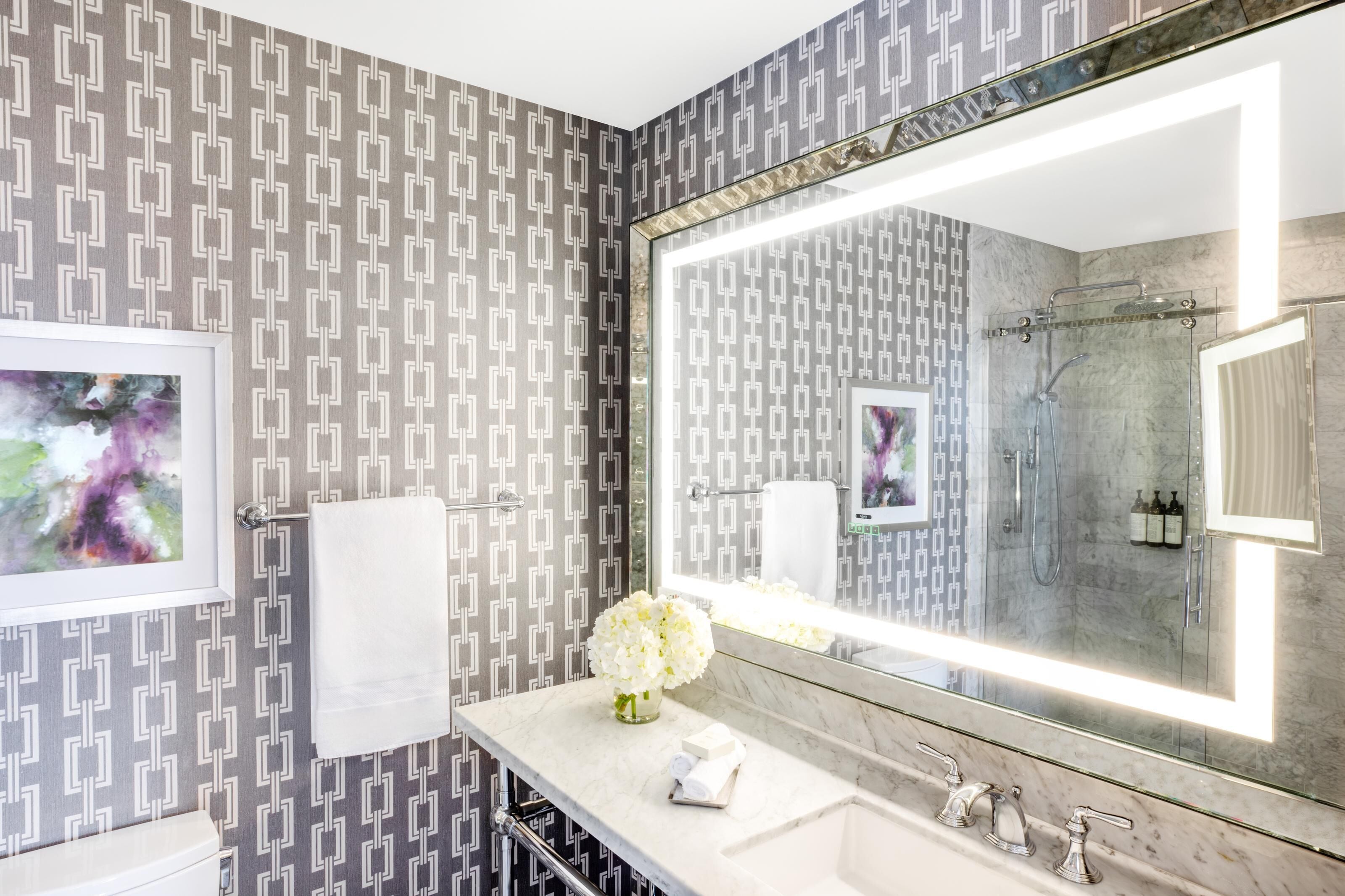 A bathroom with patterned wallpaper, a large mirror, a sink, a toilet, and a shower.